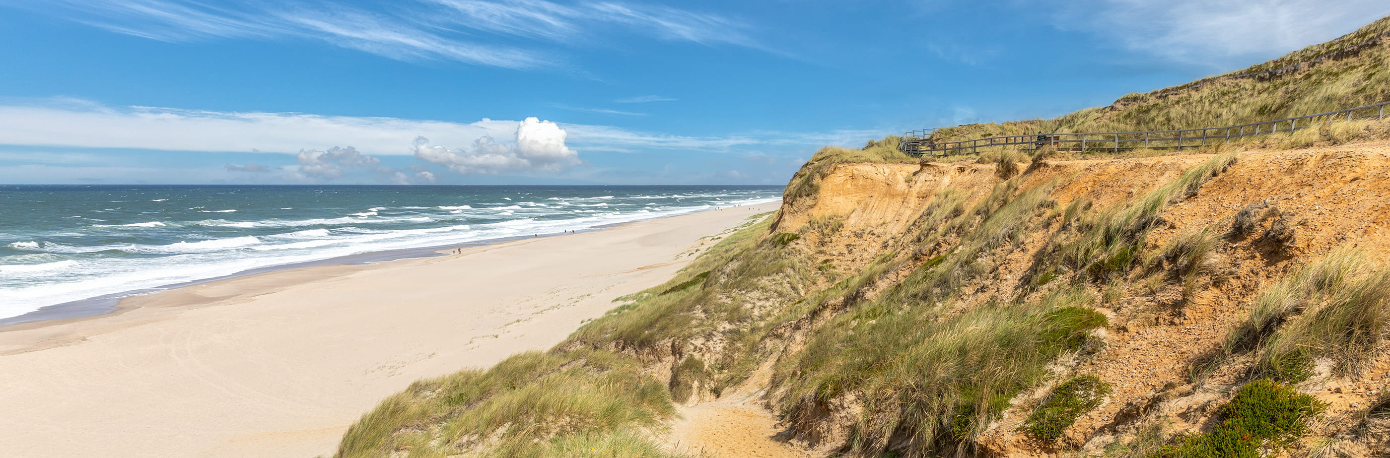 Steilküste in Kampen auf Sylt mit Dünen, Sandstrand und Nordseewellen unter blauem Himmel.